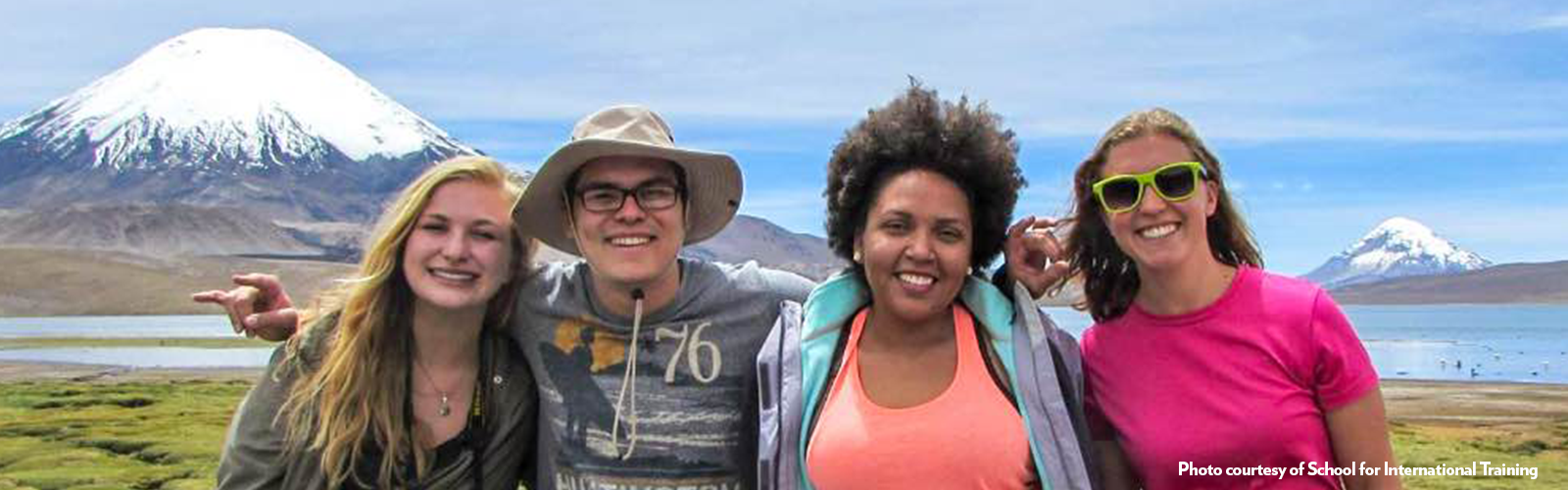 four students are standing in front of a mountain range. they are all in activewear and are smiling at the camera.